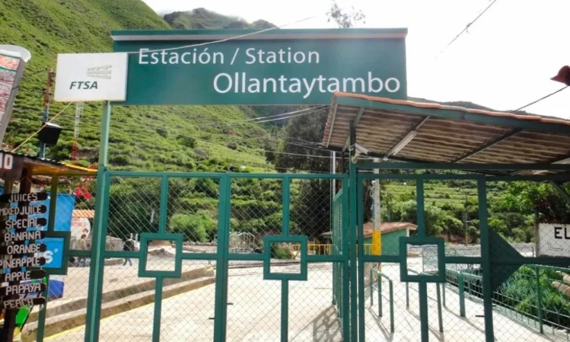 Ingreso a la Estación de Tren de Ollantaytambo Letrero verde de la Estación Ollantaytambo con rejas de ingreso y montañas verdes al fondo