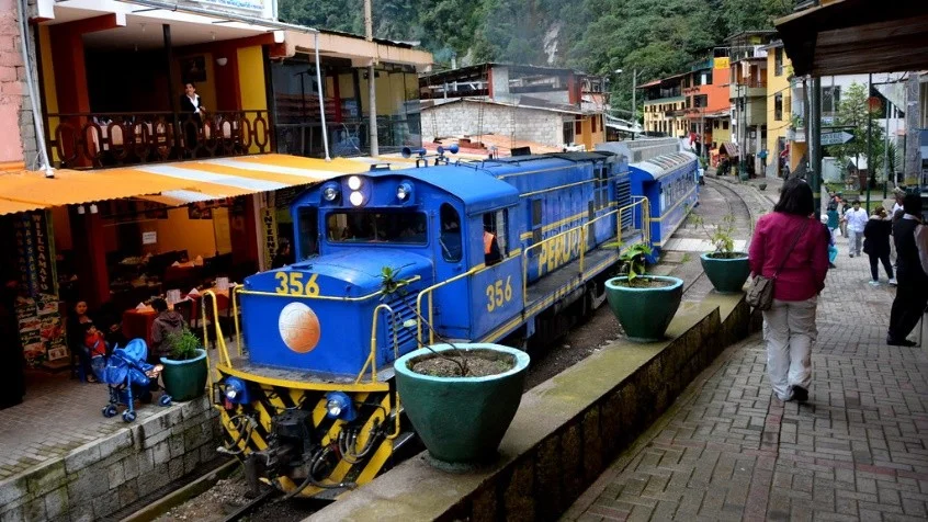 Tren de PeruRail en Aguas Calientes Tren azul de PeruRail transitando por las vías públicas de Machu Picchu Pueblo o Aguas Calientes junto a restaurantes y turistas